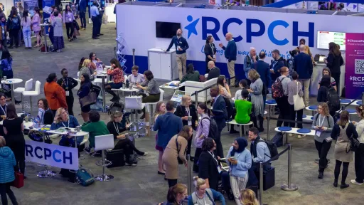 Lots of people sitting and standing in an exhibition hall, with a sign reading RCPCH