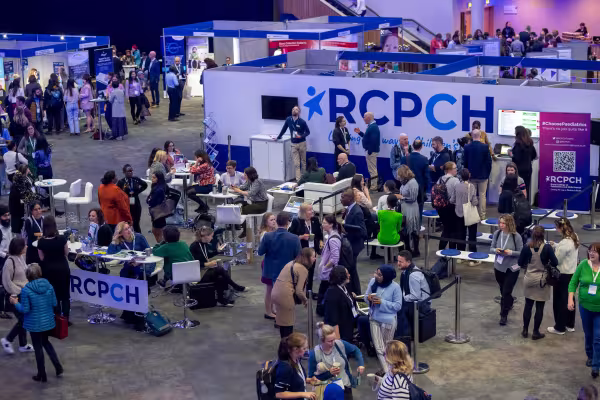 Crowd of people sitting and standing, talking to one another, in an exhibition hall; with RCPCH in large writing